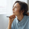 Portrait of pensive thoughtful dreaming curly adorable businesswoman looks aside pondering about right work decision can not choose reclines on fist with pen at office. Copy space for ad